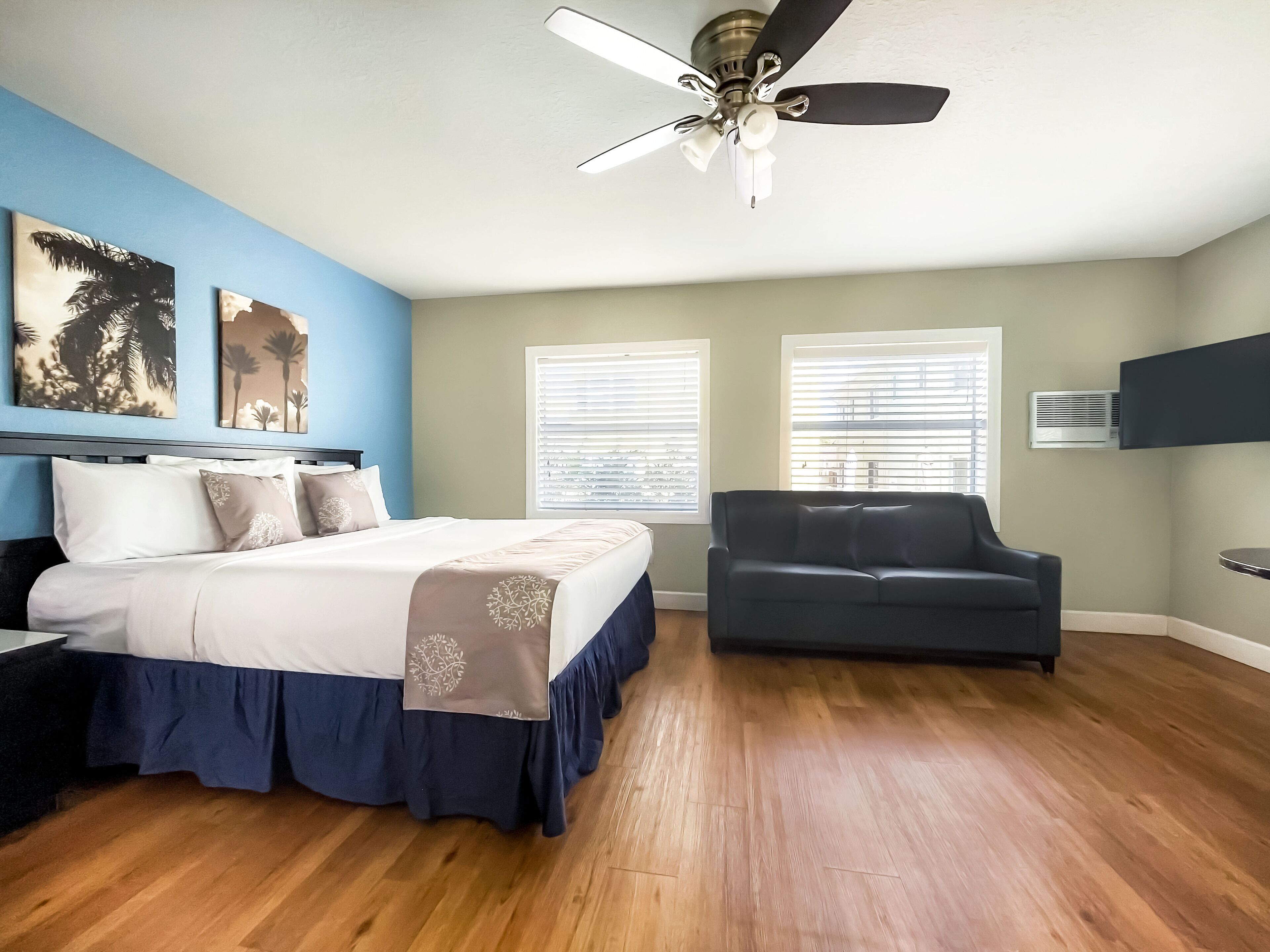 A bedroom with a bed, a dark-colored couch, two windows with blinds, a ceiling fan, and an air conditioning unit.