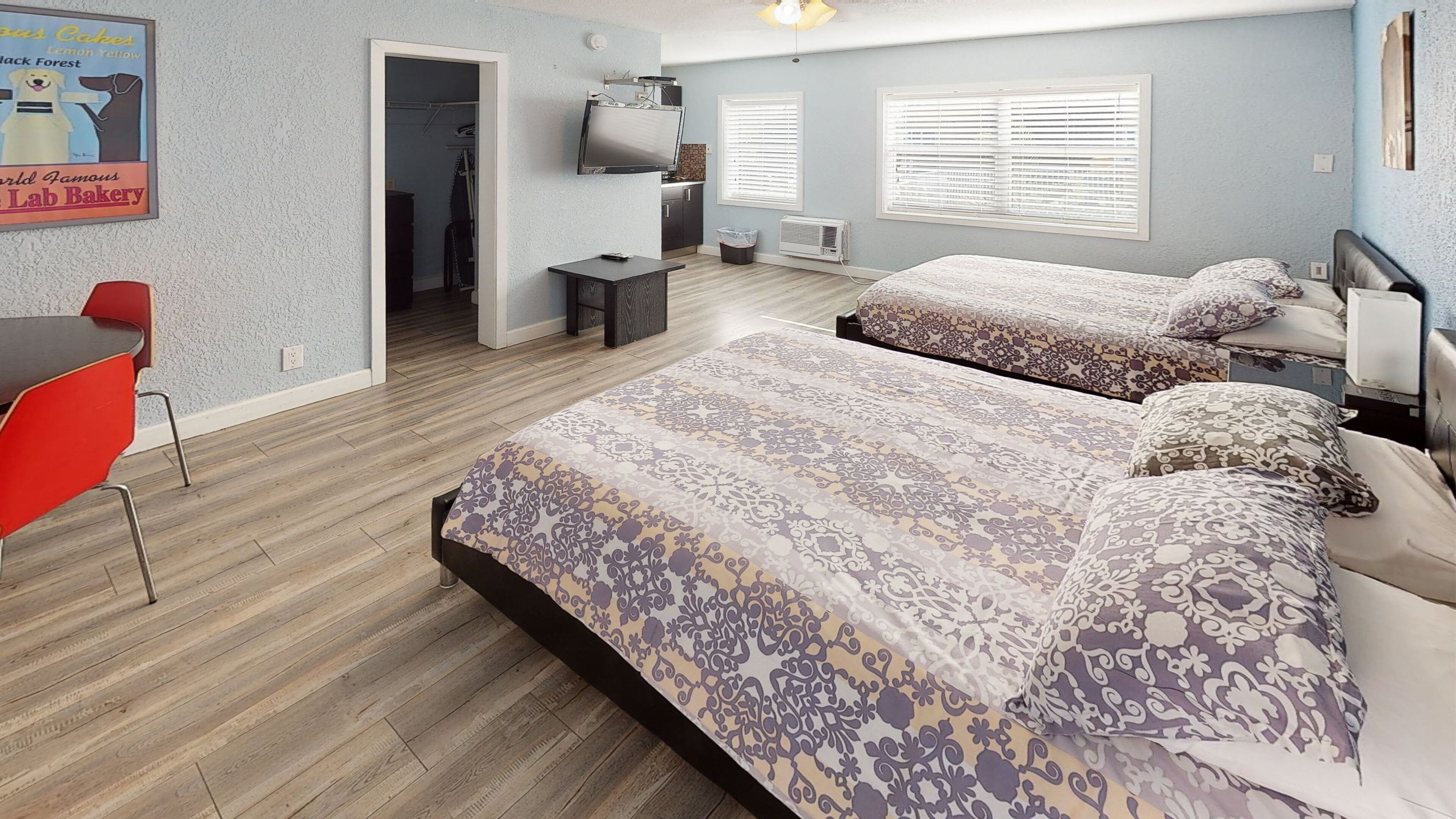 A modern bedroom with two beds, a desk, and a chair. There is a TV mounted on the wall, and a window with blinds.