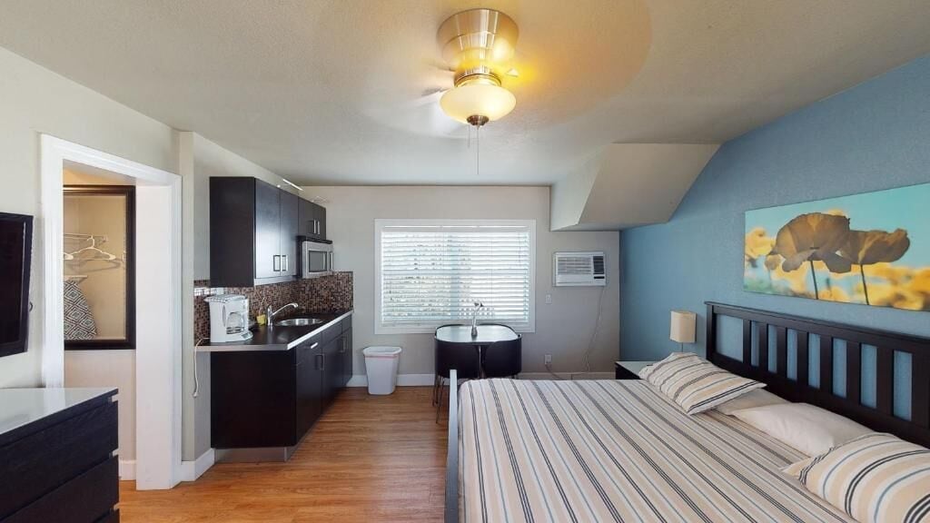 A compact bedroom with a bed, a small kitchenette, a ceiling fan, and a window with blinds.