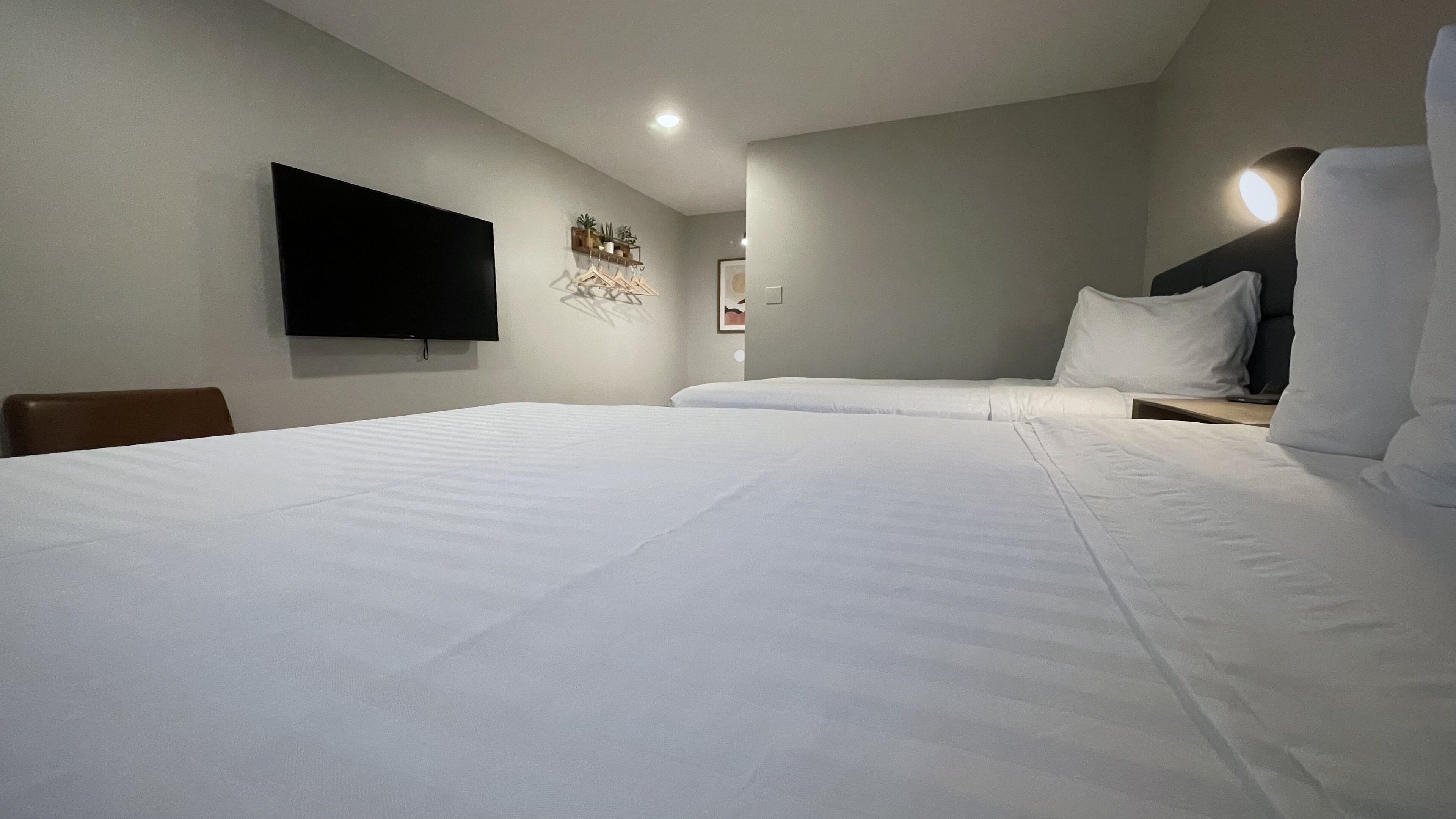 A hotel room with two beds, a flat-screen TV mounted on the wall, a bedside table with a lamp, and a small shelf with decorative items.