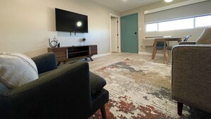 Una sala de estar moderna con un televisor de pantalla plana montado en la pared, un mueble de madera para entretenimiento, un tapete estampado y una puerta verde azulada que da a otra habitación.