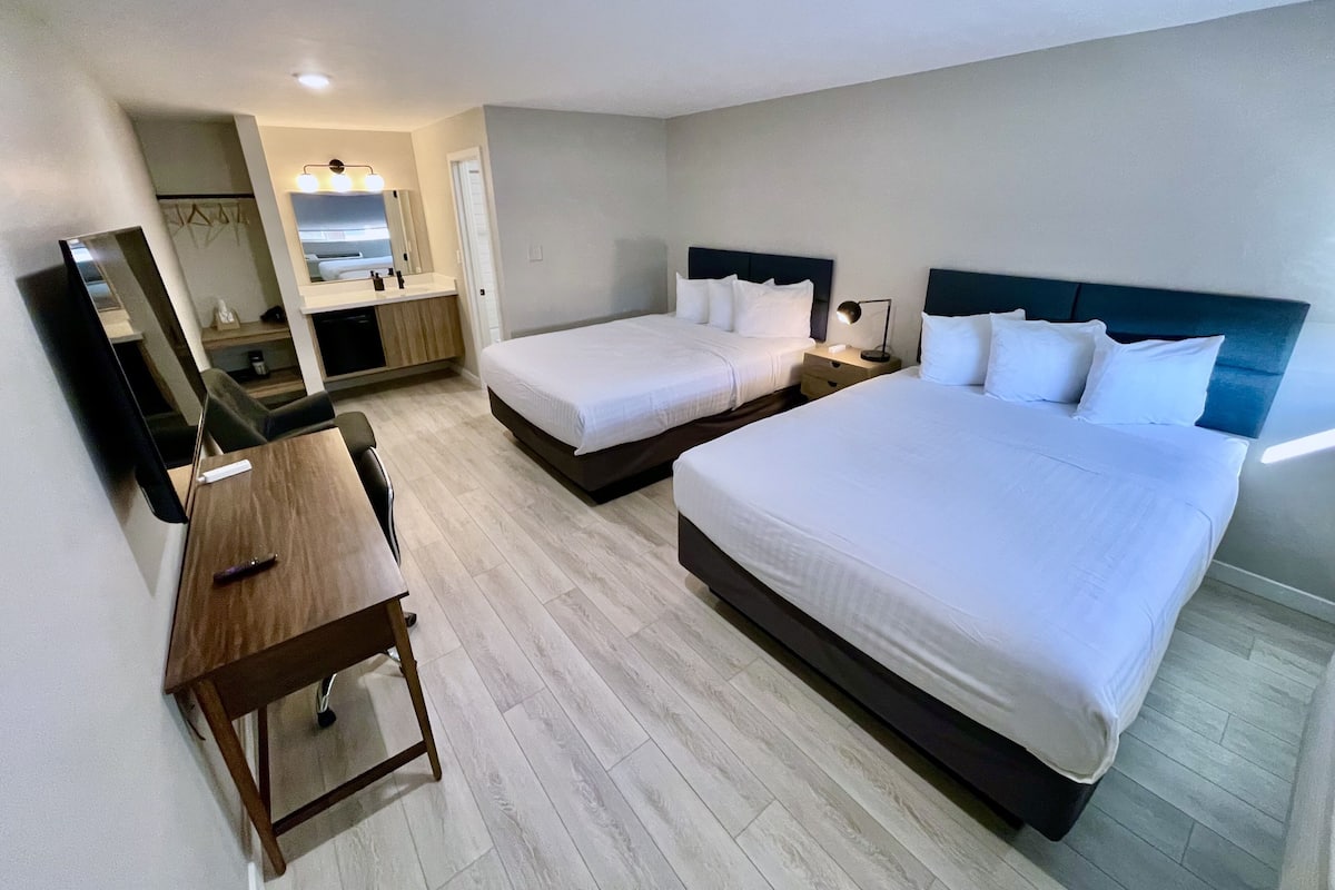 Une chambre d’hôtel avec deux lits simples, un bureau, une chaise, un miroir, une petite table et une salle de bain visible à travers une porte ouverte.
