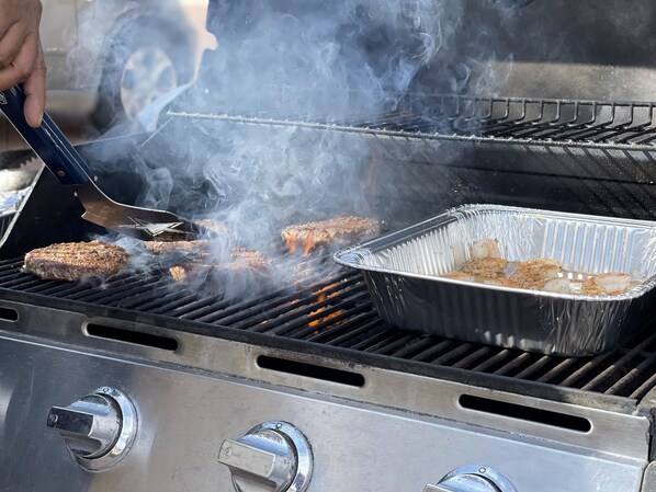 A grill with multiple burgers cooking, a pan with shrimp, and a person using tongs.