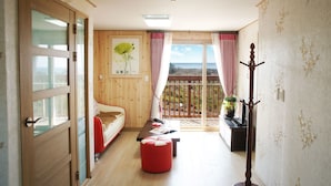 Een kamer met een houten vloer, een rode bank, een klein rood poufje, een houten kapstok en een balkon met uitzicht.