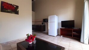 Een woonkamer met een salontafel, een televisie op een houten dressoir, een koelkast en een keuken in de achtergrond.