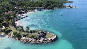 An aerial view of a coastal area with a rocky island, clear turquoise waters, and a beach with umbrellas. - The Cove Suites at Blue Waters Resort and Spa (St. John's)