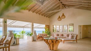 A spacious room with a large window overlooking the sea, a wooden ceiling, and a central seating area with a glass coffee table. - The Cove Suites at Blue Waters Resort and Spa (St. John's)