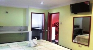Habitació d'hotel amb un llit, un banyet petit, un televisor muntat a la paret i una porta vermella que dona a una altra habitació.