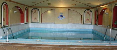 A large indoor swimming pool with a tiled floor and walls, surrounded by red-framed arches and a decorative ceiling.