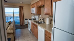 Cozinha com armários de madeira, geladeira branca, micro-ondas e lava-louças. Há uma janela com vista para a praia.