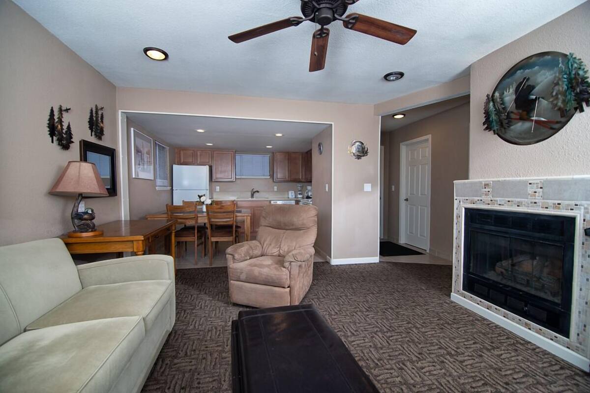 Una sala de estar con un sofá color beige, un sillón reclinable, chimenea, ventilador de techo y una cocina al fondo.
