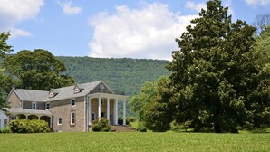 A large stone house with a wrap-around porch and columns, surrounded by lush green fields and trees. - Mountain Cove Farms Resort (Chickamauga)