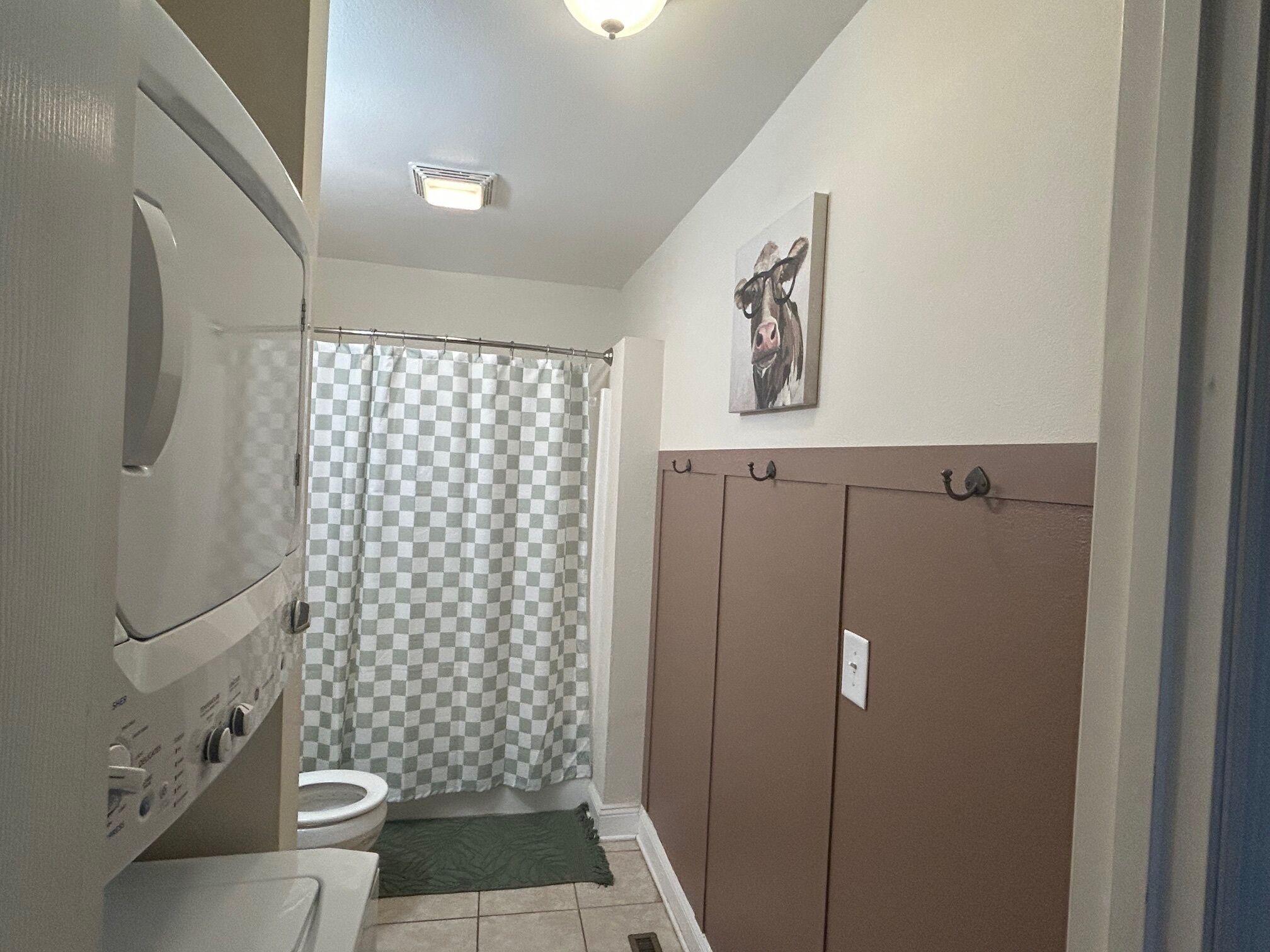 A compact bathroom with a toilet, a checkered shower curtain, a green mat, and a wall-mounted towel rack.