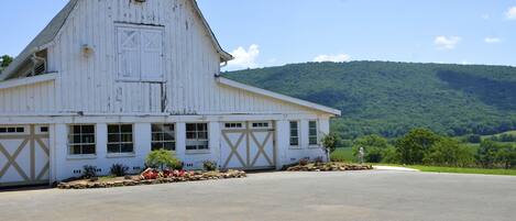A white barn with a large garage door, surrounded by a paved driveway and a landscaped area with flowers and shrubs.
