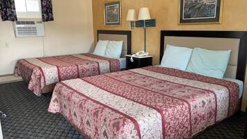 A hotel room with two beds, each with a patterned bedspread, a bedside table with a lamp, and a wall-mounted air conditioner.