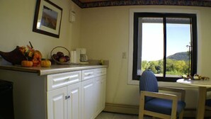 A kitchen with white cabinets, a dining table with a blue chair, a window with a view of hills, and a framed picture on the wall. - Appalachian Motel (Vernon)