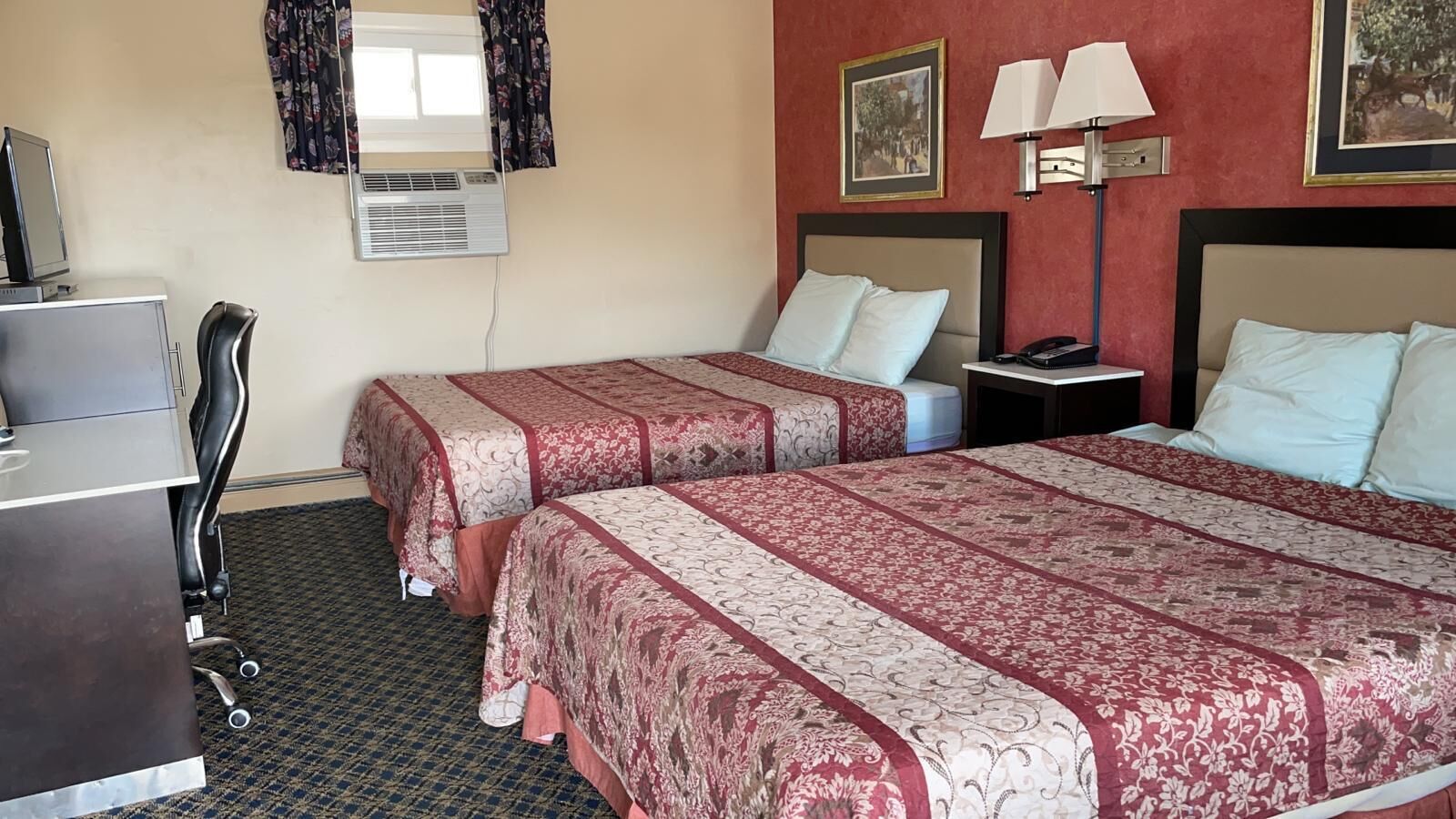Habitación de hotel con dos camas, un escritorio, una silla, un equipo de aire acondicionado y una mesita de noche con lámpara.