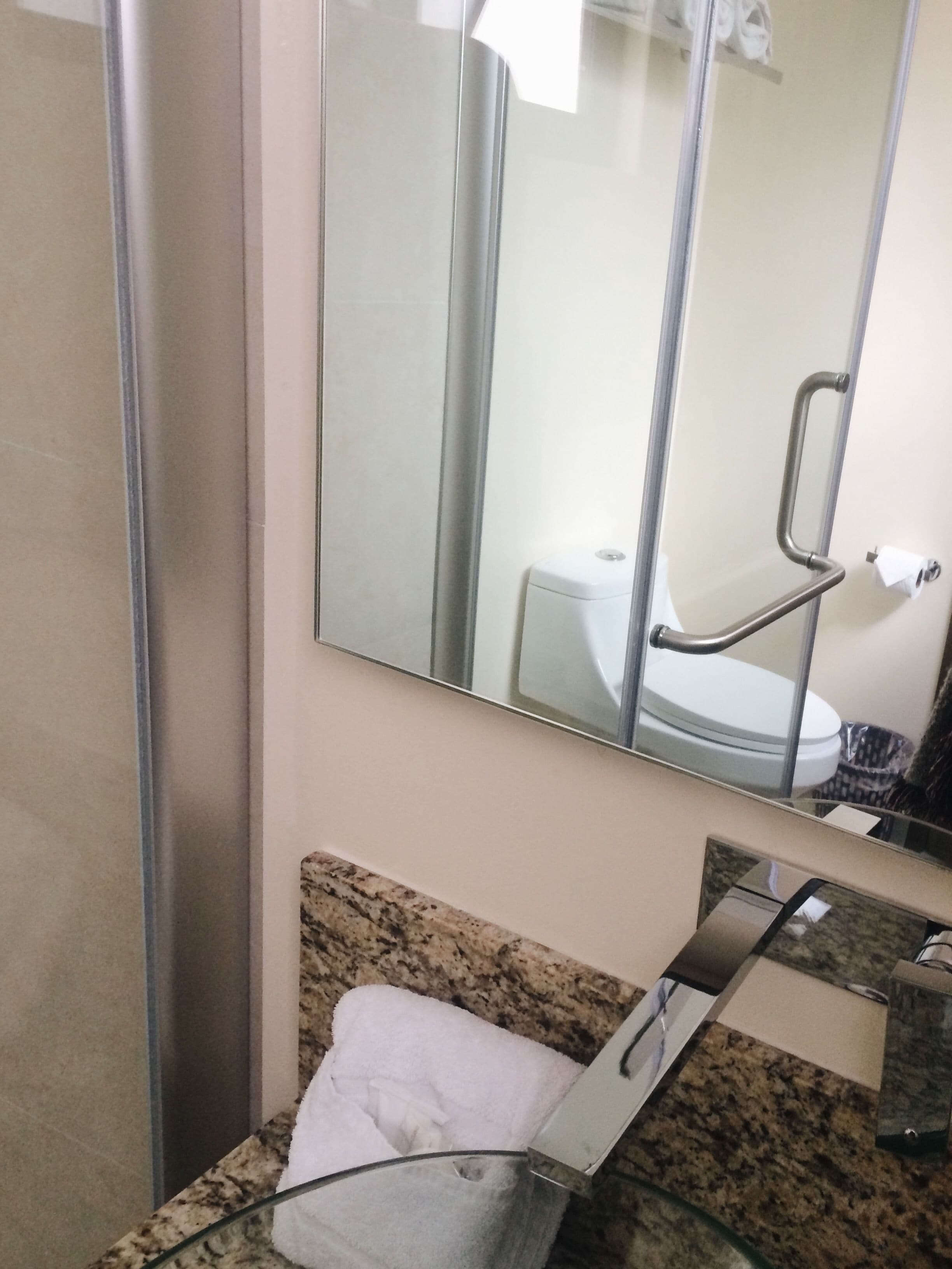 Un baño con encimera de granito, cabina de ducha con mampara de vidrio y un inodoro que se ve reflejado en el espejo.