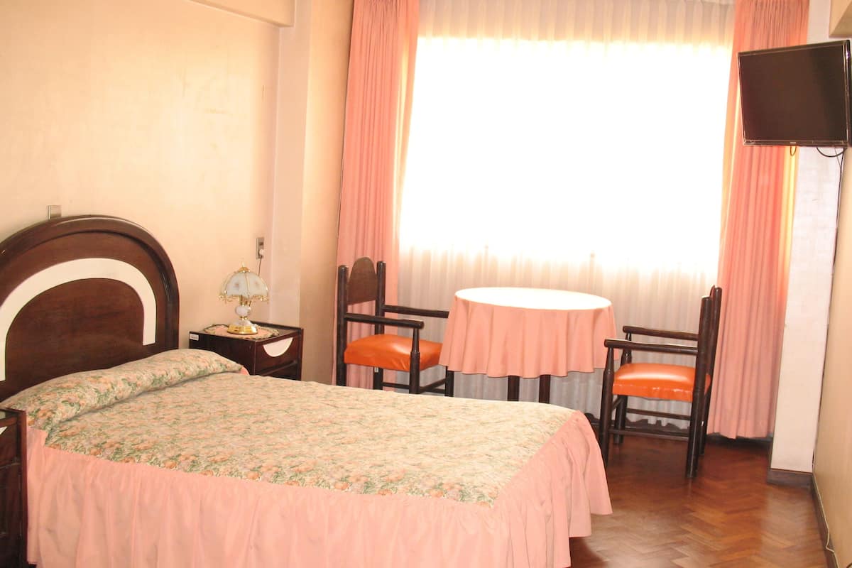 Un dormitorio con cama, mesita de noche, una mesa con dos sillas, televisor y ventana con cortinas.