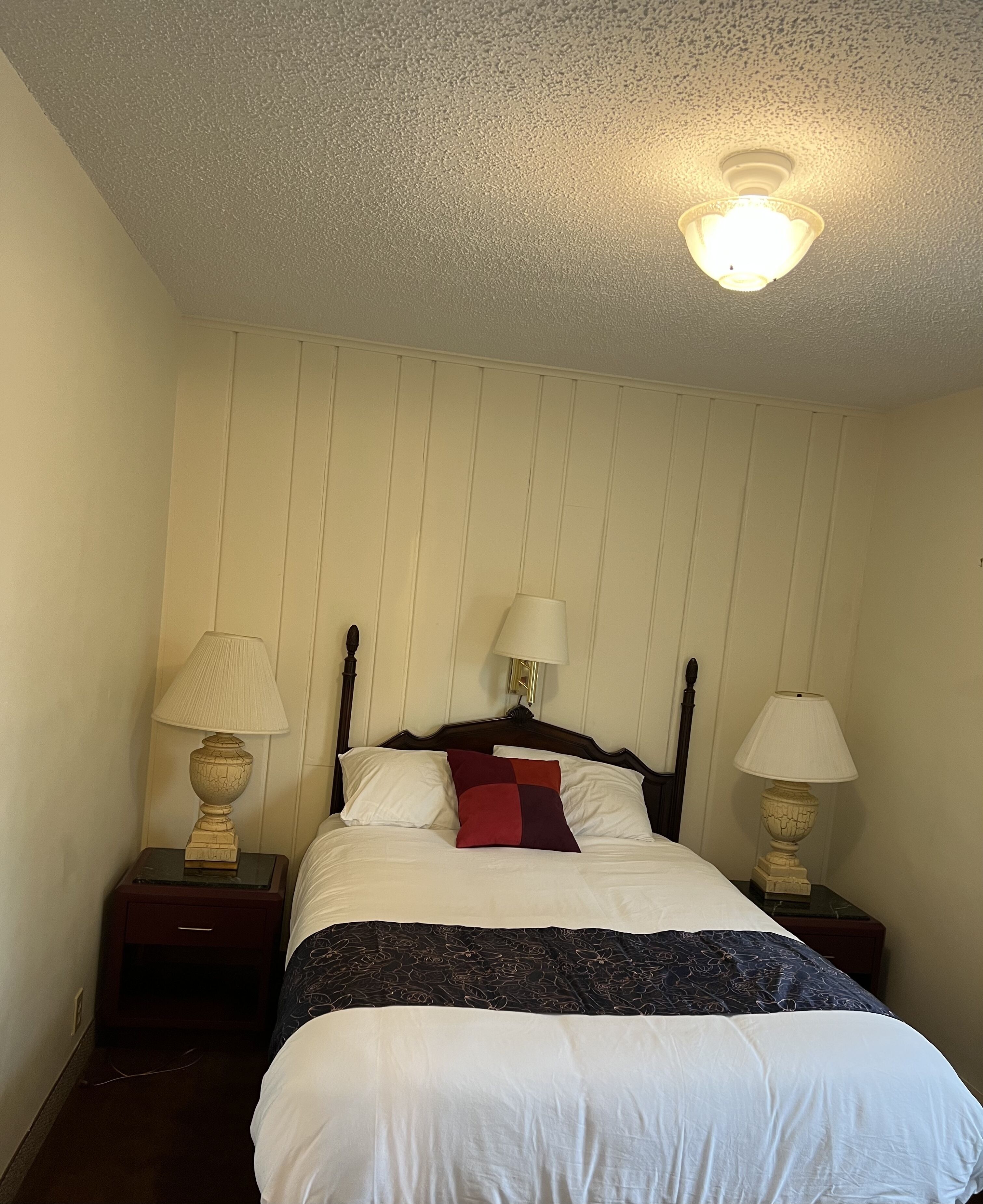 A neatly made bed with white and dark blue bedding, two bedside lamps, and a ceiling light fixture.
