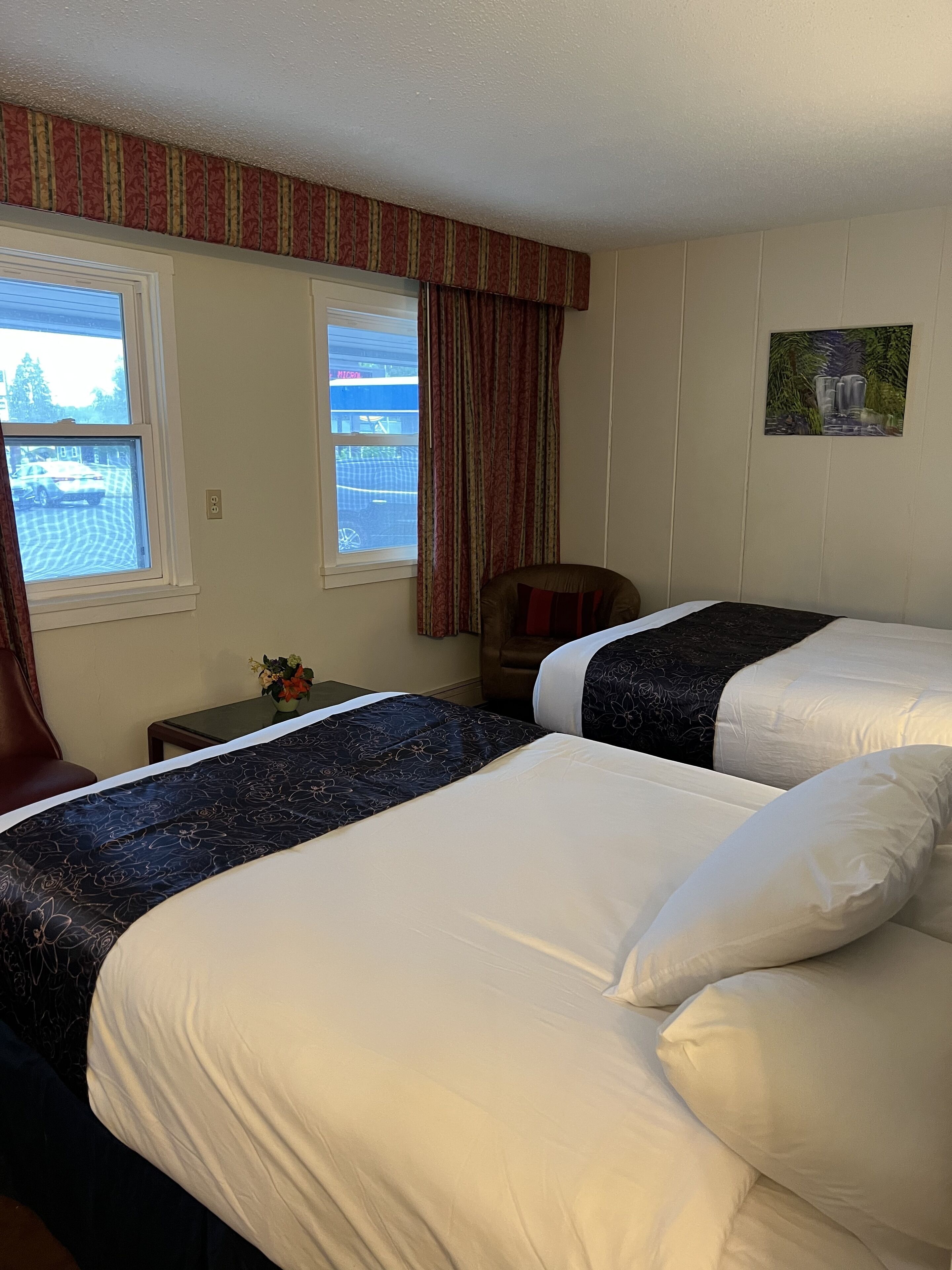 A hotel room with two beds, a window with a view of a parking lot, a small table with a vase of flowers, and a painting on the wall.