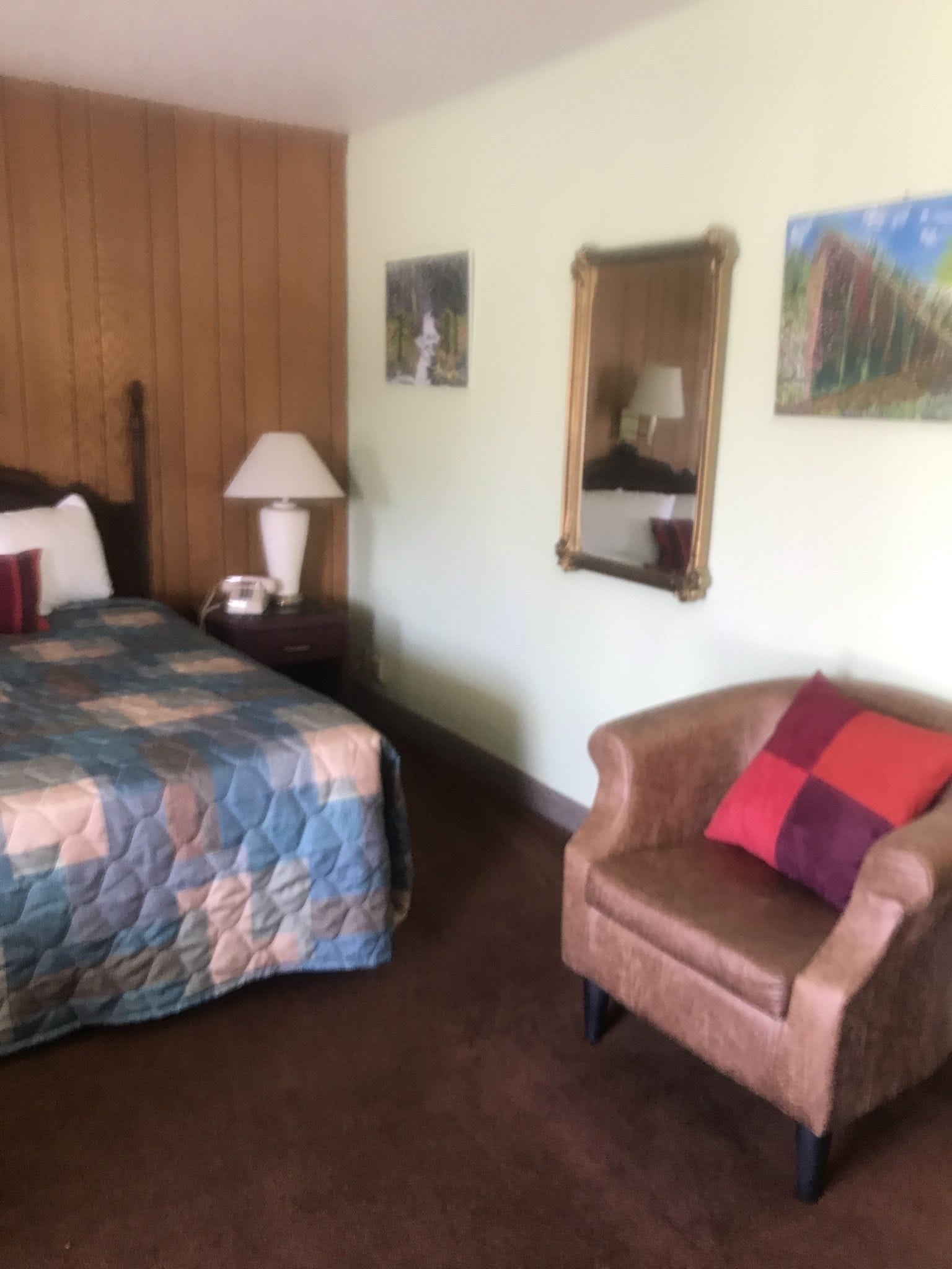 A bedroom with a bed, a chair, a nightstand, a lamp, a mirror, and two paintings on the wall.