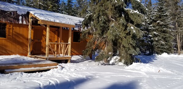 A wooden cabin with a porch, surrounded by snow-covered trees. - Lonesome Dove Guest Ranch (Kalispell)