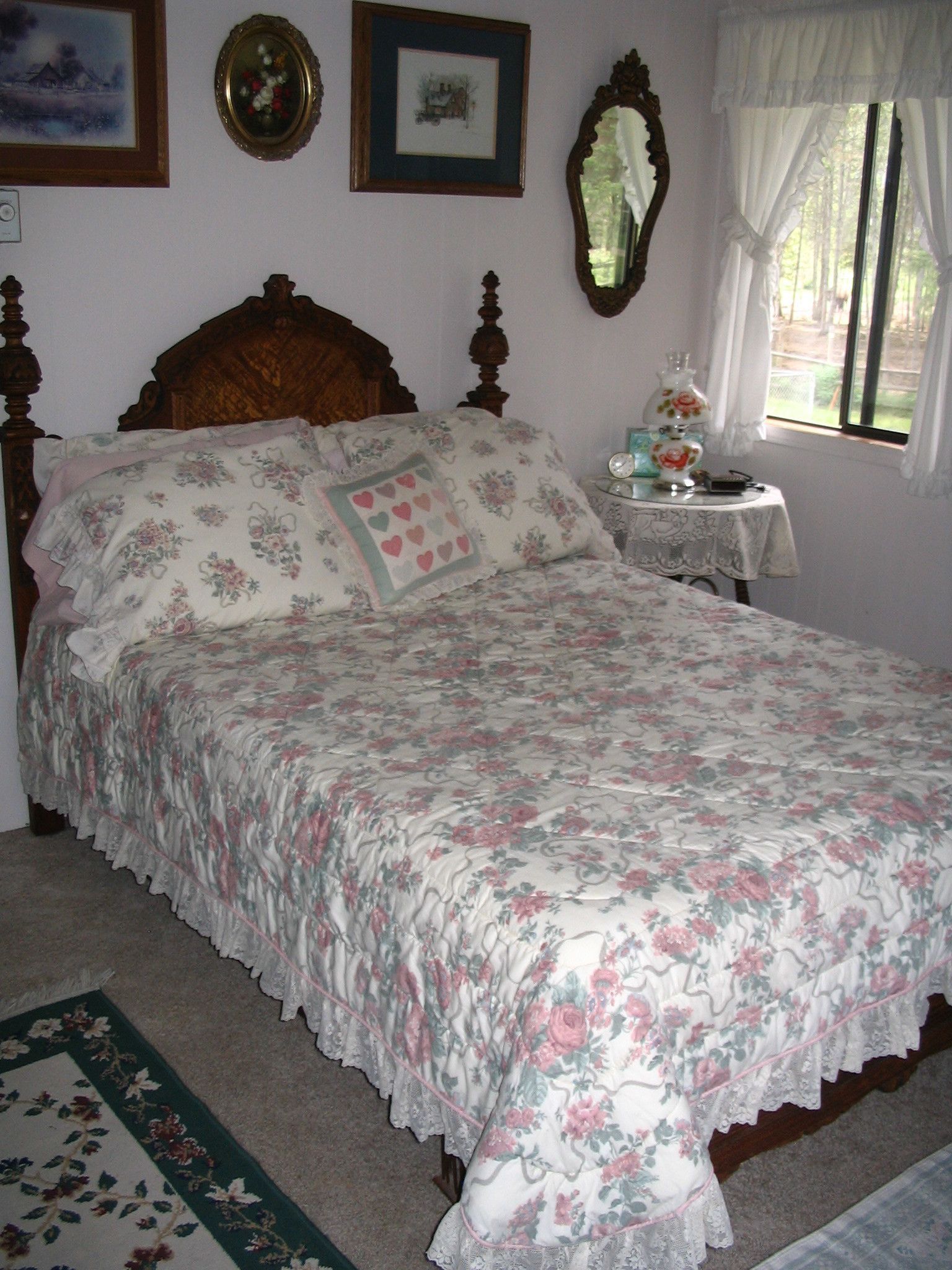 A bedroom with a floral bedspread, a wooden headboard, a bedside table with a lamp, and a window with curtains.