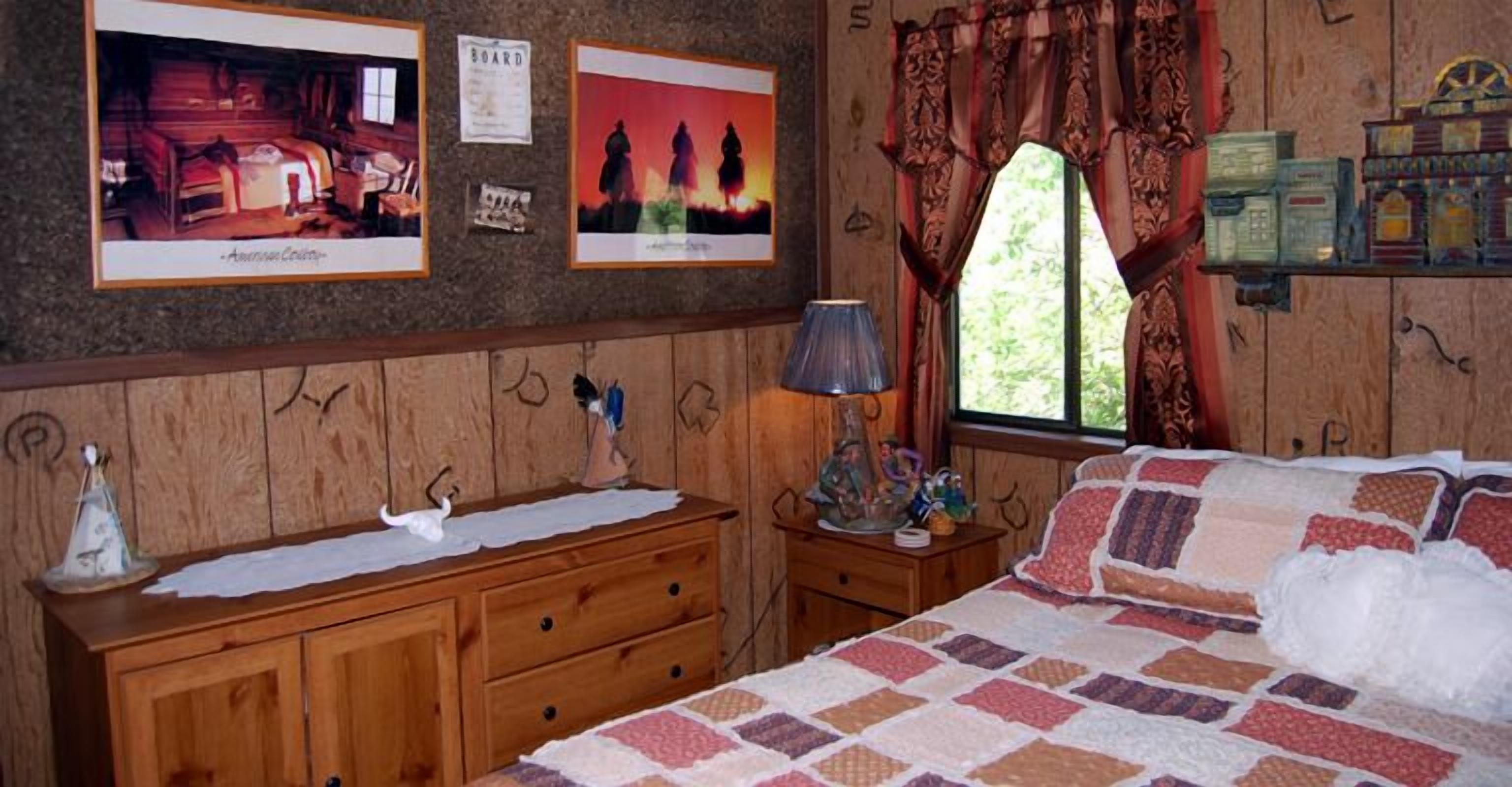 A bedroom with a wooden dresser, a bed with a quilt, a nightstand with a lamp, and a window with curtains.