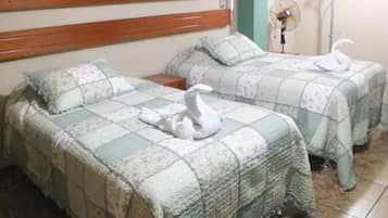 Une chambre avec deux lits, chacun recouvert d’une couvre-lit à motifs floraux et agrémenté d’une décoration en forme de cygne blanc. On y trouve une tête de lit en bois, un ventilateur fixé au mur et un sol carrelé.