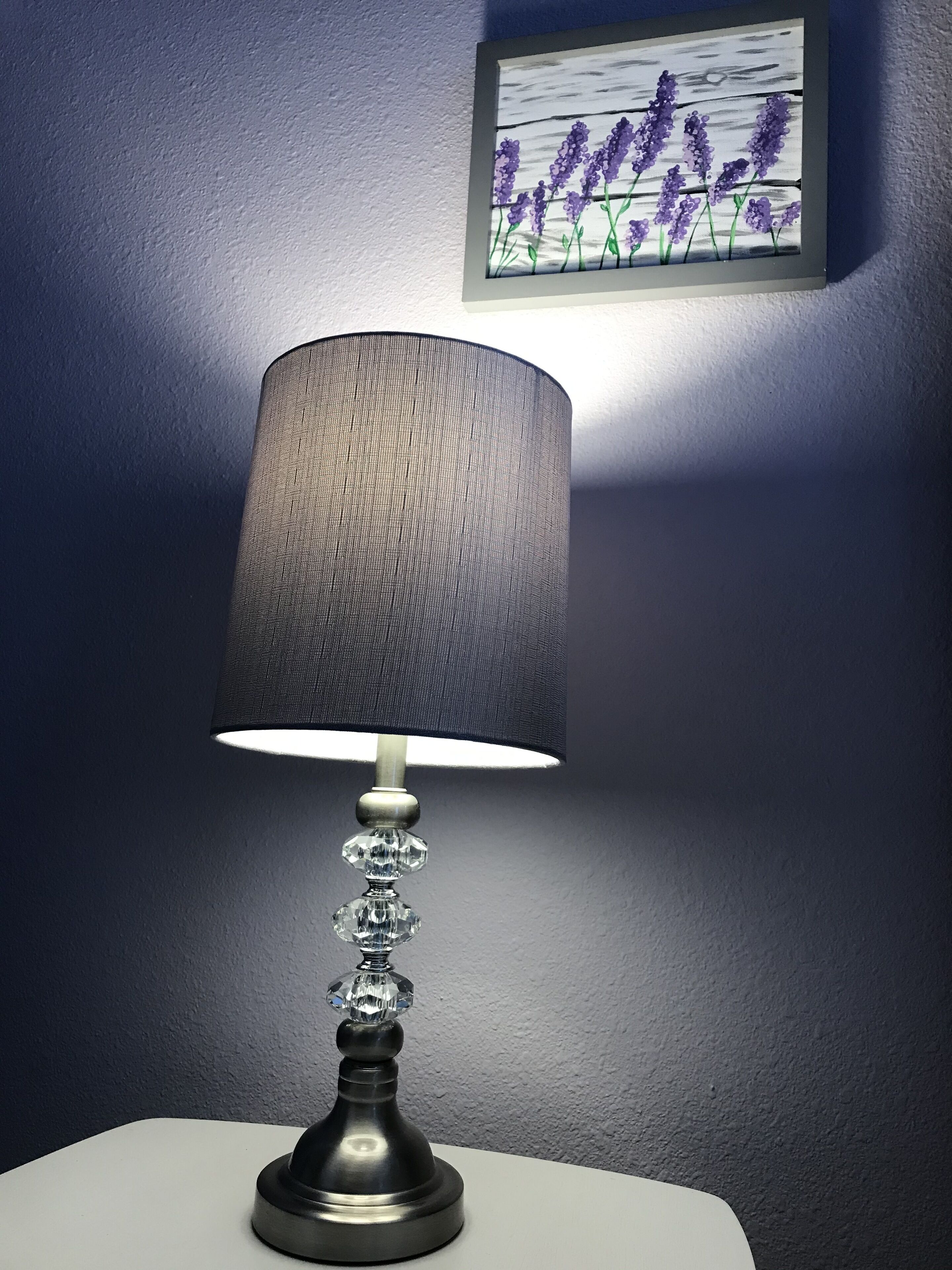 A table lamp with a textured shade and crystal accents on a white table against a dark wall.