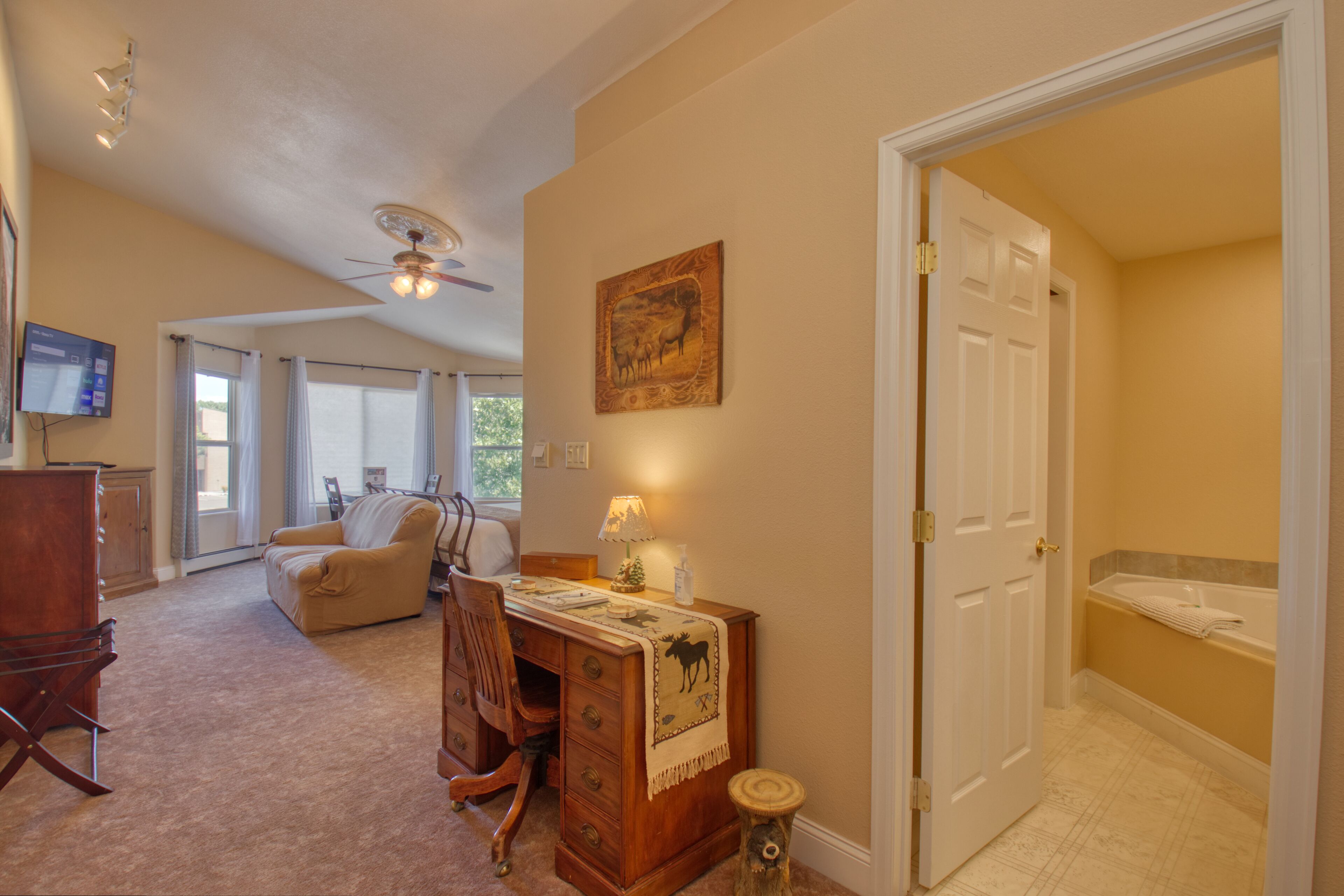 A spacious living room with a large window, a ceiling fan, a wooden desk, and a cozy seating area.