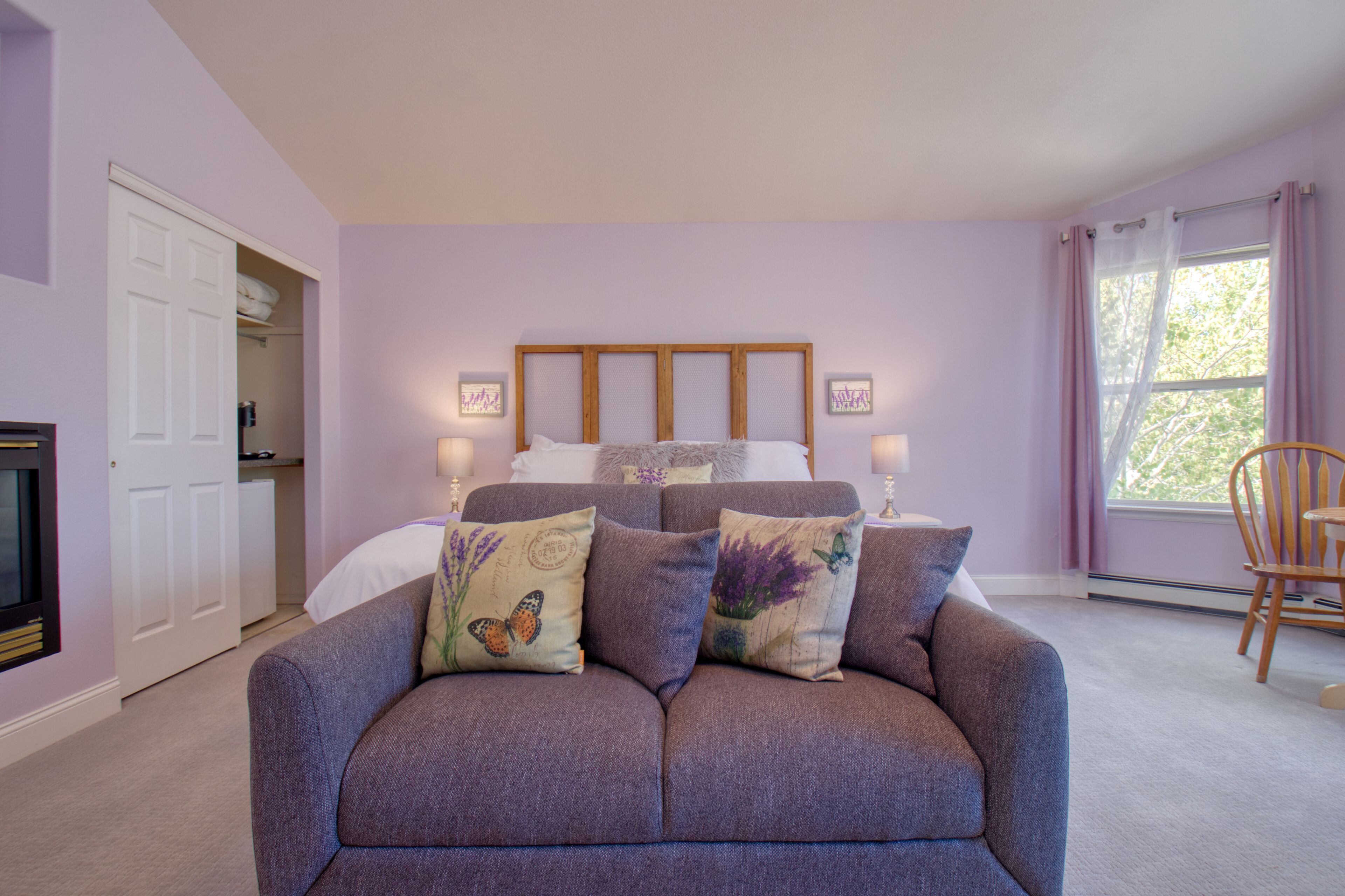 Quarto com cama, sofá cinza, cabeceira de madeira, duas luminárias de cabeceira, janela com cortinas e uma cadeira.