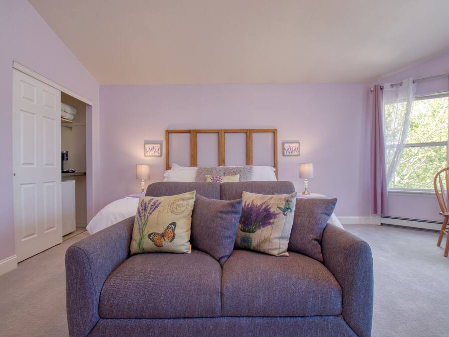 Quarto com cama, sofá cinza, cabeceira de madeira, duas luminárias de cabeceira, janela com cortinas e uma cadeira.