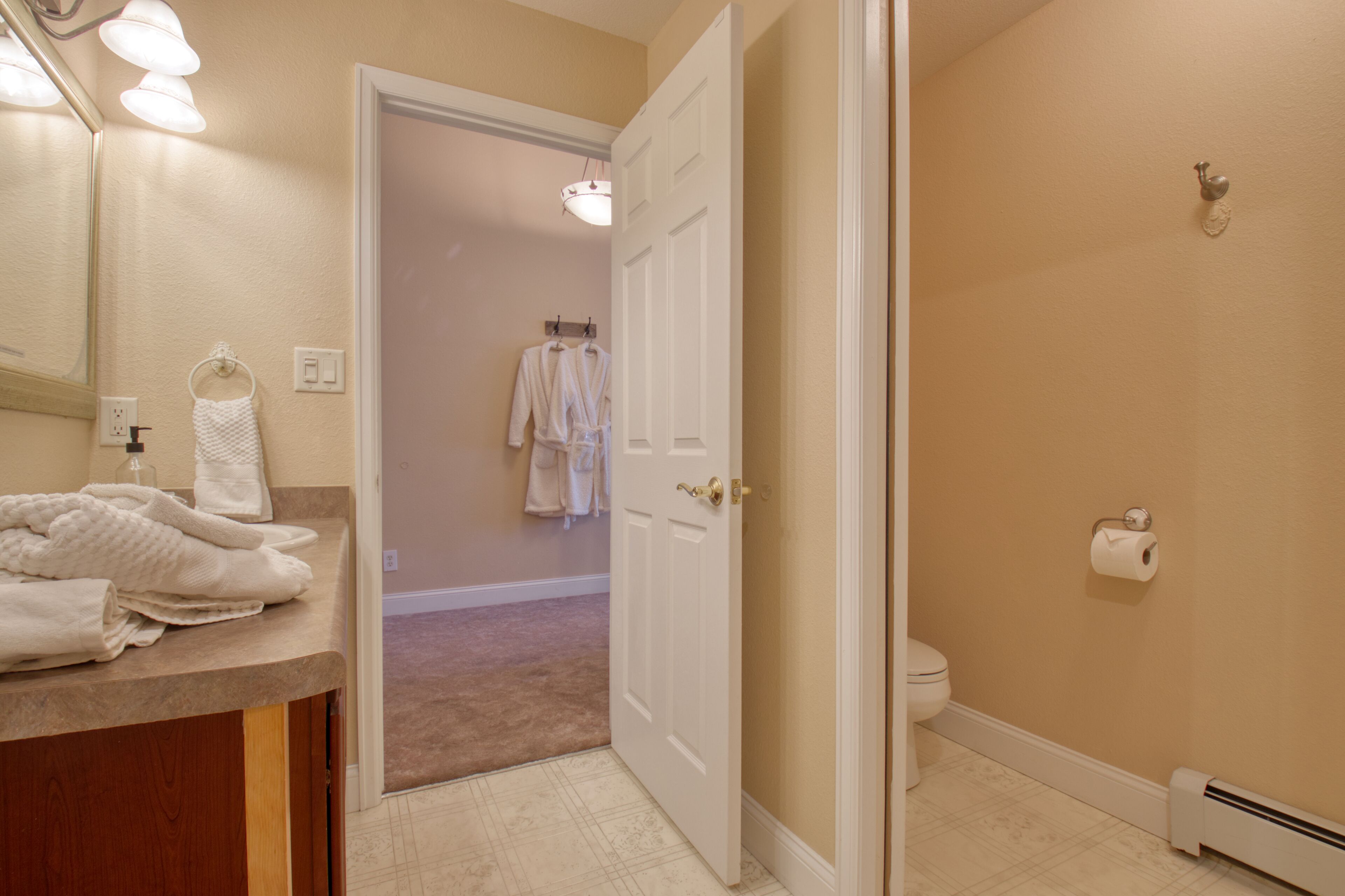 A bathroom with a beige wall, a white toilet, a towel rack with towels, and a mirror with lights.