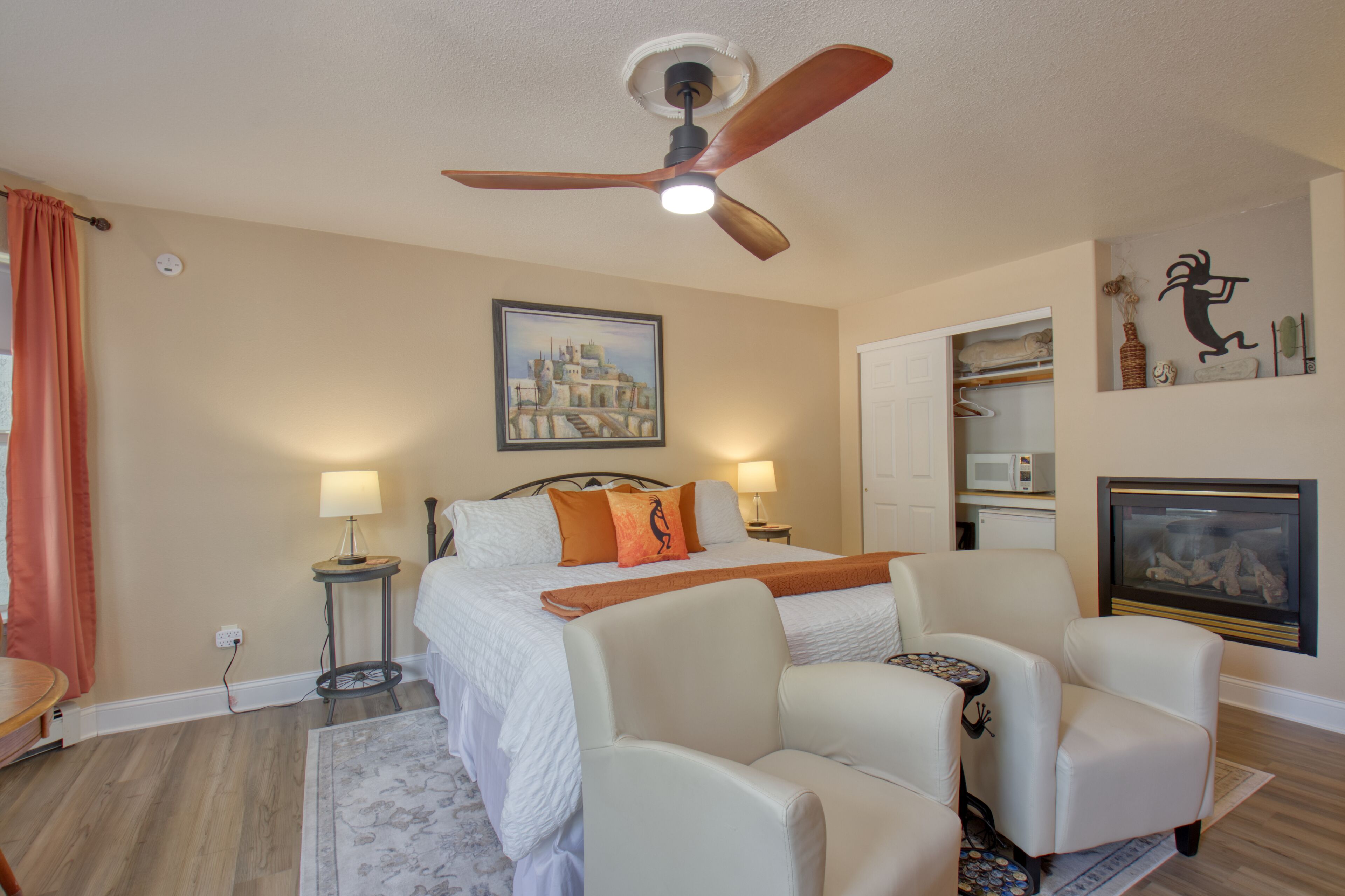 A hotel room with a bed, two bedside lamps, a ceiling fan, a painting on the wall, a microwave, and a television.