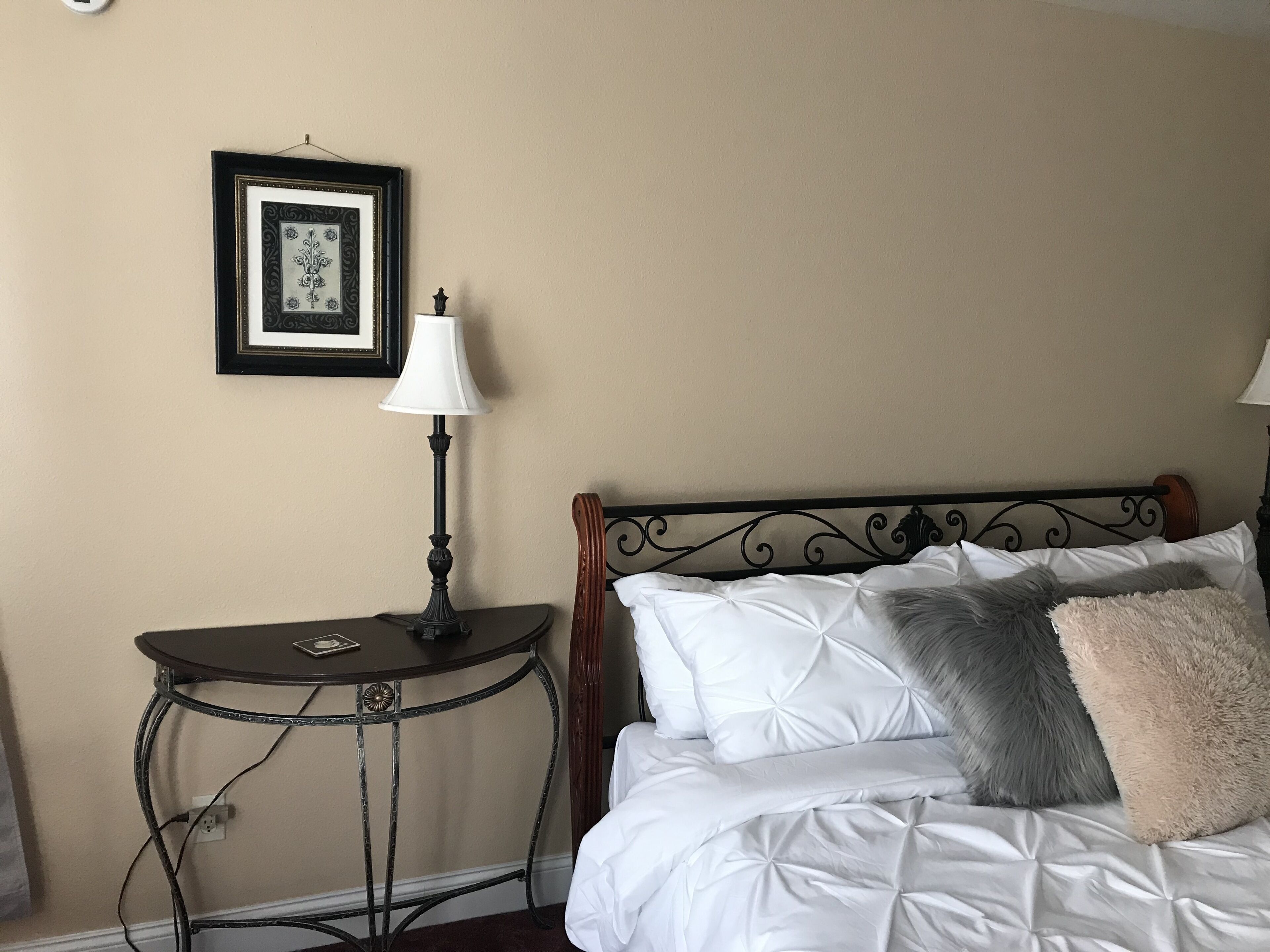Ein ordentlich bezogenes Bett mit weißer Bettwäsche und einem grauen Kopfkissen, ein kleiner Tisch mit einer Lampe und ein gerahmtes Bild an der Wand.