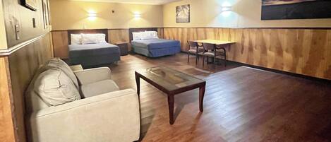 A hotel room with a sofa, two single beds, a small table, and a dining area with chairs.