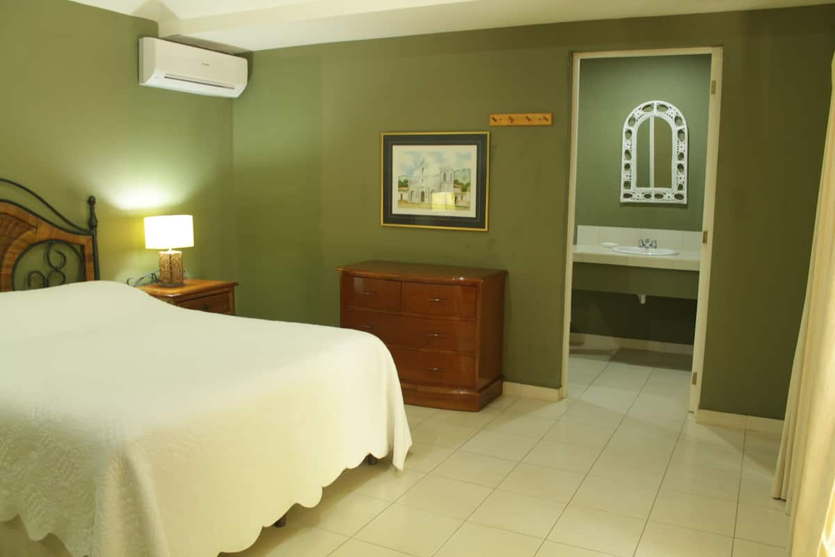 A bedroom with a bed, dresser, nightstand, lamp, framed picture, and a bathroom with a sink and mirror.