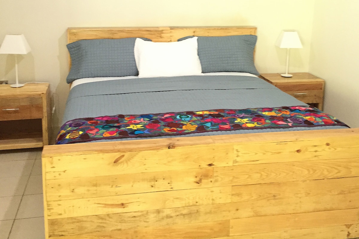 A wooden bed with a colorful quilt, two bedside tables with lamps, and a wall-mounted air conditioner.
