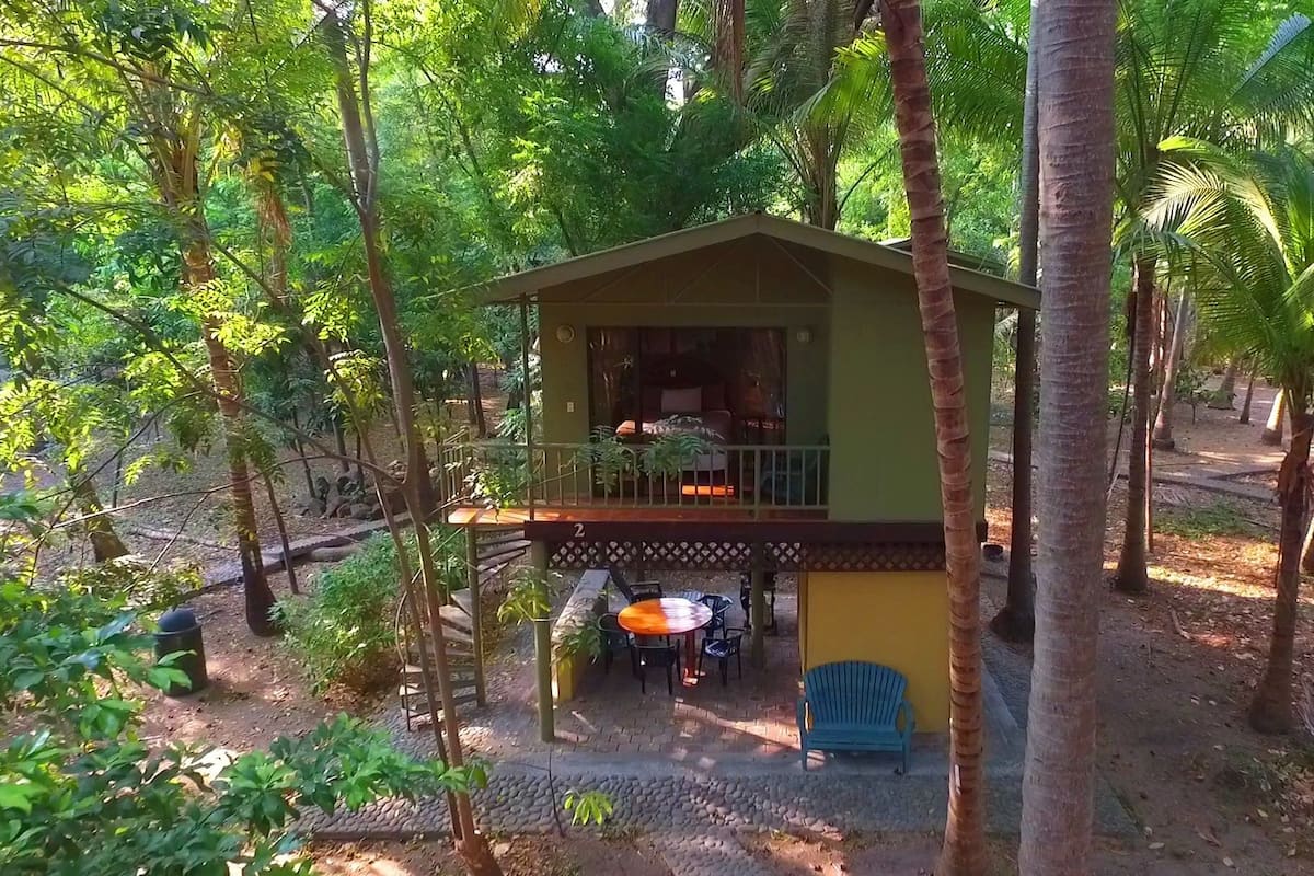 A small, single-story wooden cabin with a balcony, surrounded by lush greenery and trees.
