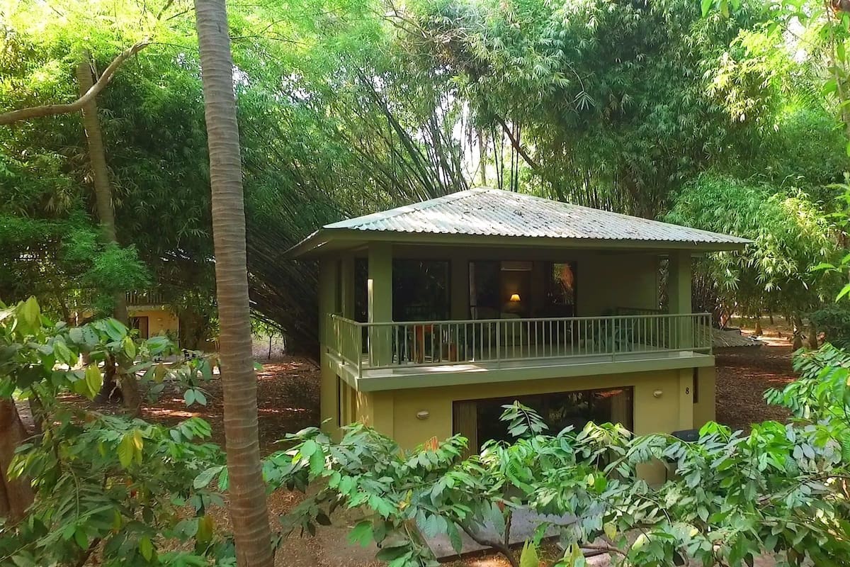 A small, single-story building with a balcony, surrounded by dense greenery.