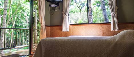 A room with a bed, a TV mounted on the wall, and a view of trees through the window.