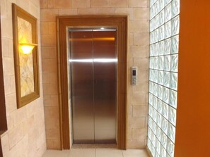 An elevator with a metallic finish, a wall-mounted light fixture, and a tiled wall with a geometric pattern. - Hotel Real Guanacaste (San Pedro Sula)
