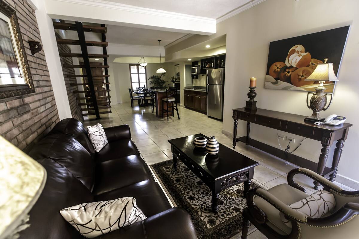 A living room with a black leather sofa, a coffee table with decorative items, a patterned rug, and a dining area in the background.