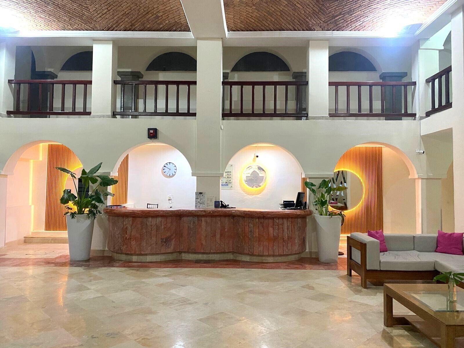 Un espace d’accueil comprenant un comptoir de réception, deux grands plantes en pot, une horloge et un coin salon avec un canapé et une table basse.