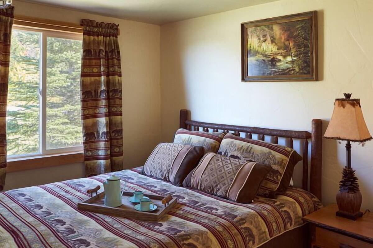 A bedroom with a bed, a painting, a window with curtains, and a nightstand with a lamp.