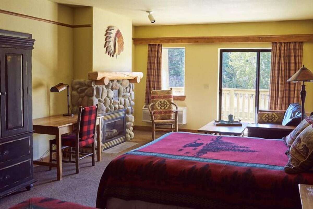 A cozy room with a fireplace, a bed with a red blanket, a desk, and a chair.