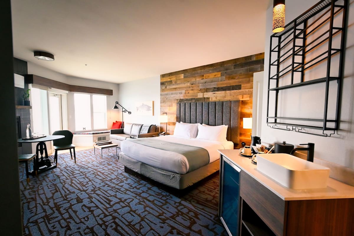 A hotel room with a large bed, a desk with a chair, a built-in TV, a bathroom with a sink and mirror, and a wooden accent wall.