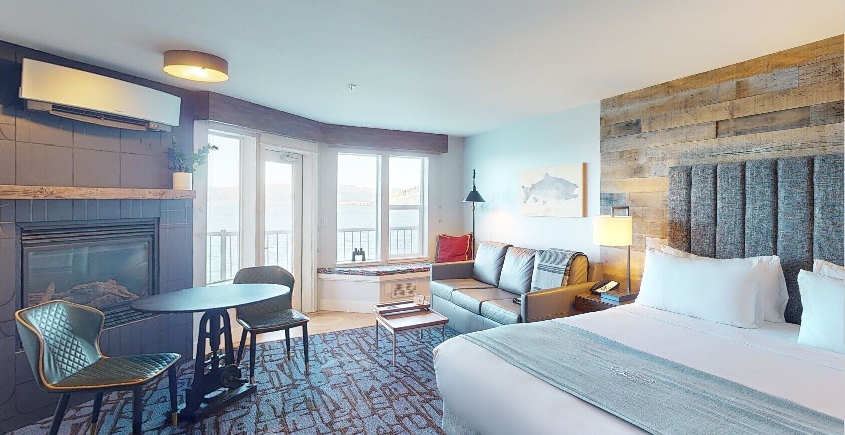 Habitación de hotel con cama, sofá, mesa de comedor y chimenea. Se ve un puente a través de la ventana.